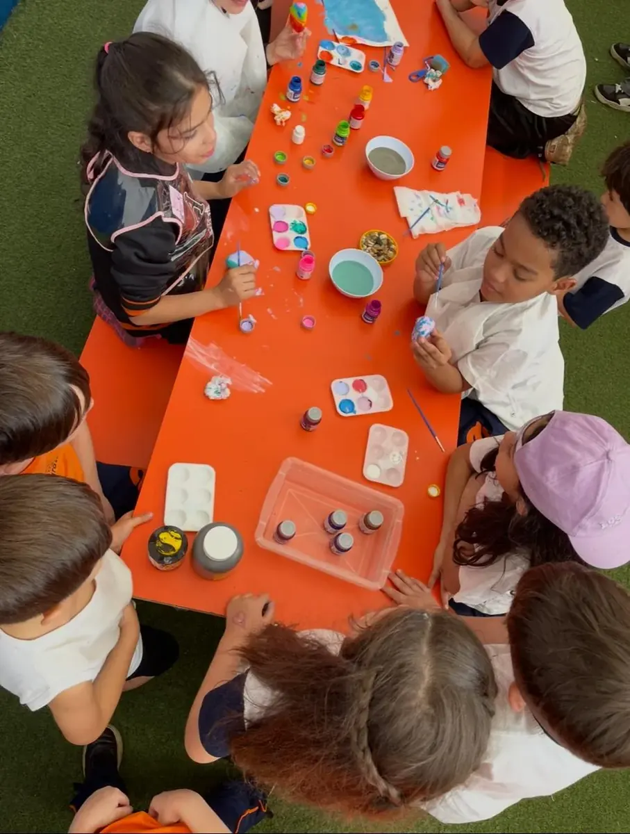 Turma do Fundamental Anos Iniciais em aula de Artes, pintando na mesa da área externa com tintas e paletas
