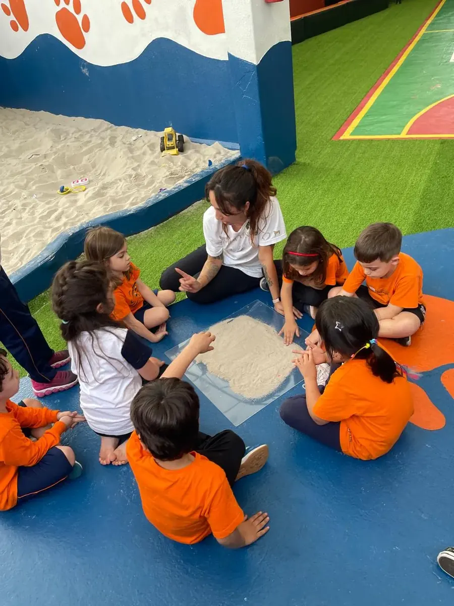 Turma da Educação Infantil sentada em roda no pátio, explorando areia numa bandeja com a professora