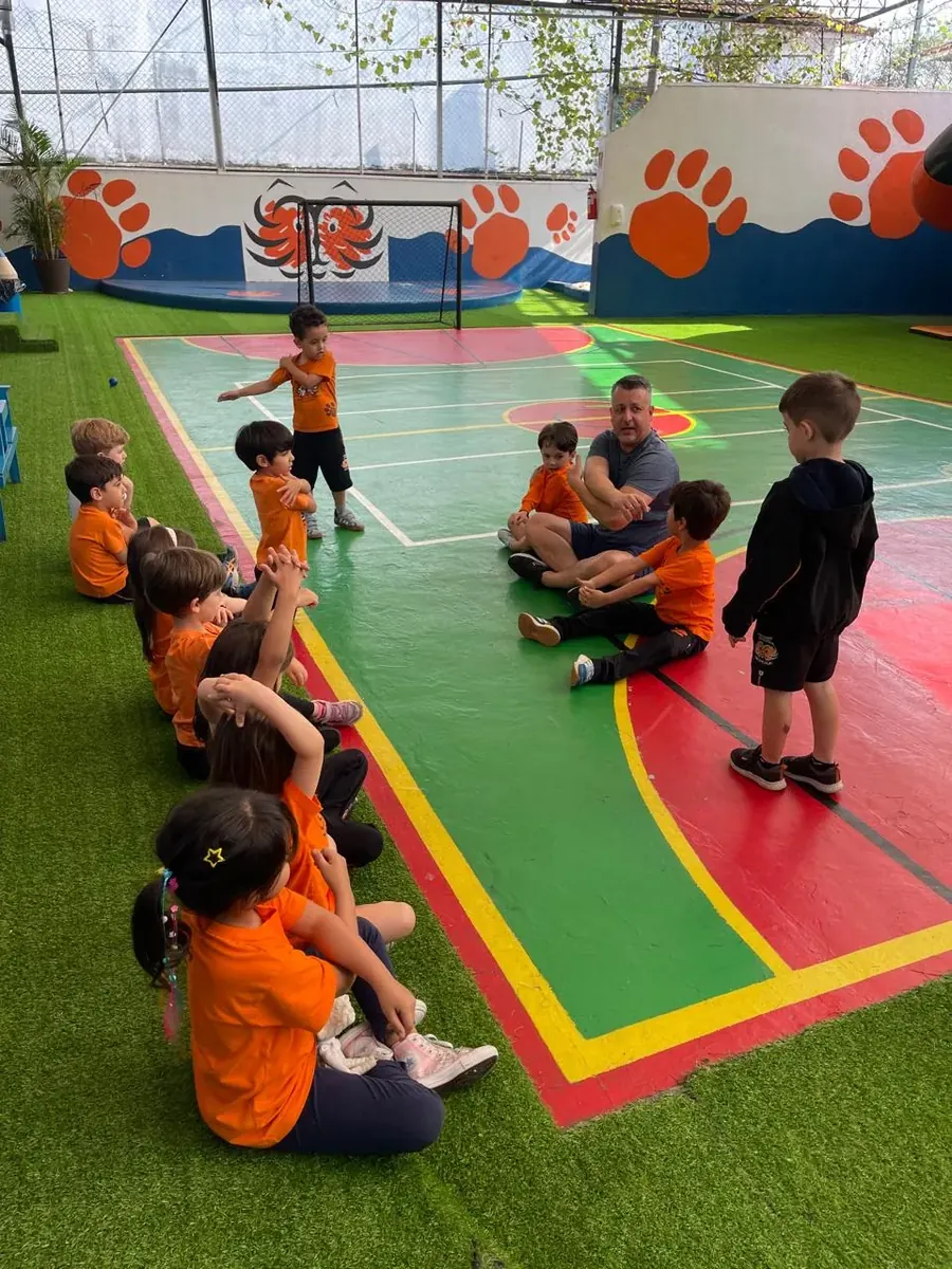 Turma da Educação Infantil sentada em roda na quadra, na aula de Educação Física