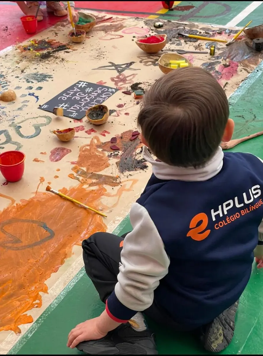 Aluno da Educação Infantil de uniforme HPLUS pintando um painel coletivo no chão, na oficina sensorial de arte