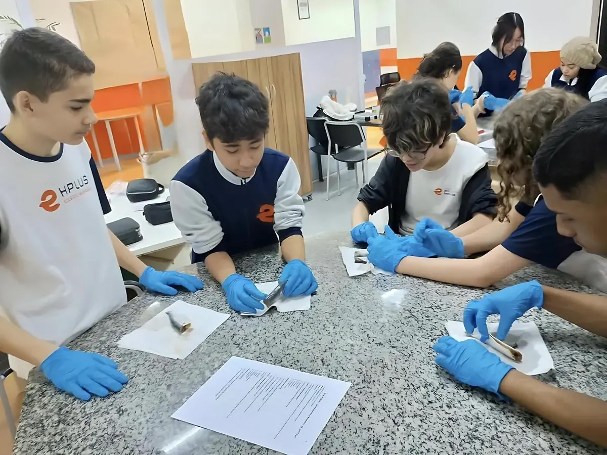 Turma do COLÉGIO HPLUS em aula prática de laboratório, de luvas, com o roteiro da atividade sobre a bancada