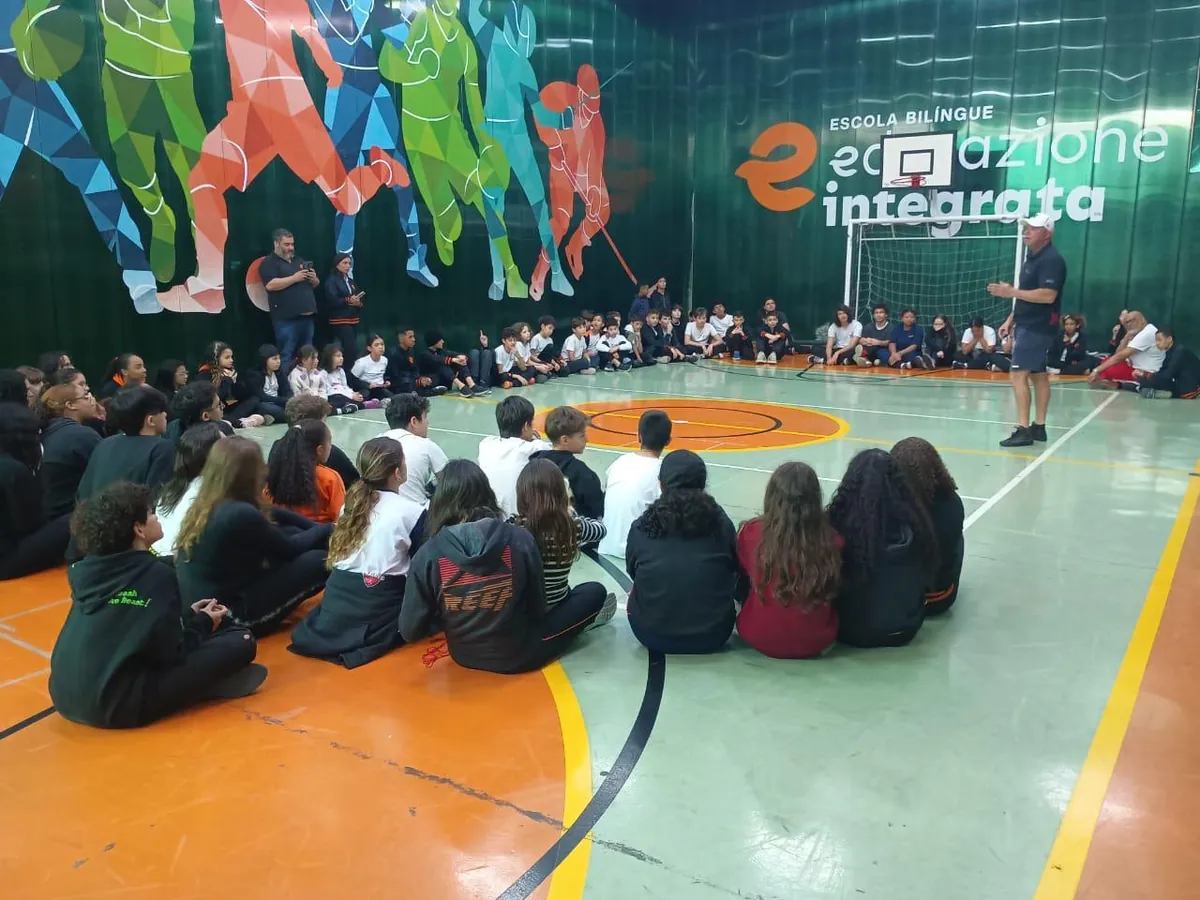 Alunos reunidos em roda na quadra poliesportiva coberta da Unidade II, com o mural esportivo ao fundo